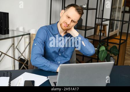 Frazzle guy using laptop for remote working, a guy tired and burnout, holding back of the head and feels ache sitting at the desk in modern office with eyes closed. Overwork concept Stock Photo