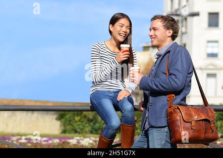 Friends talking drinking coffee on lunch break outdoor in the spring sun on city street. Happy Multiracial couple workers enjoying cafe date together Stock Photo