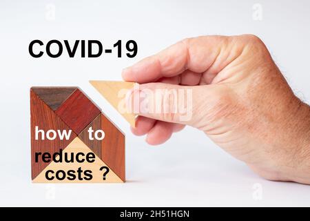 The missing building block is inserted into a tangram puzzle. The text is written on the white background: CORVIT-19 and the words how to reduce costs Stock Photo