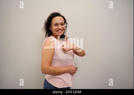 Portrait of African happy woman smiling after getting antiviral vaccine. Vaccinated woman showing thumb up and her arm with bandage after receiving im Stock Photo