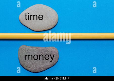 The words time and money are written on natural smooth stones separated by a yellow pencil. The background is isolated in blue and has a lot of space Stock Photo
