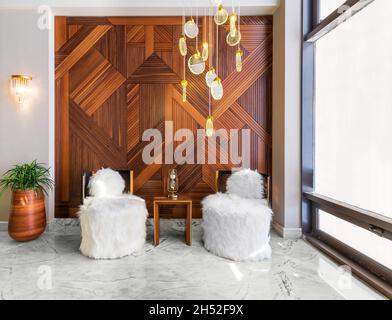 Two modern white feather armless chairs, with small wooden modern table in the middle, and wooden tall planter with green bushes, in a hall with decorated wood cladding wall, and white marble floor Stock Photo