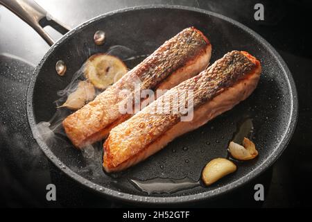 Two slices of fresh salmon fillet with a crispy fried skin in a black pan with garlic, thyme and lemon on the stove, cooking a delicious seafood meal, Stock Photo