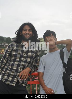 Two male friends standing together in outside with looking at camera. A long haired young guy smiling and other one covering his face with showing rock sign with his hand Stock Photo