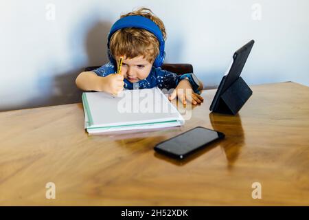 three year old boy at home sitting at a table with tablet and headphones doing homework. childhood and technology concept Stock Photo