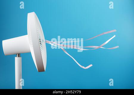 The electric fan is white with pink ribbons fluttering in the wind on a blue background. Free space, minimalistic style. Stock Photo