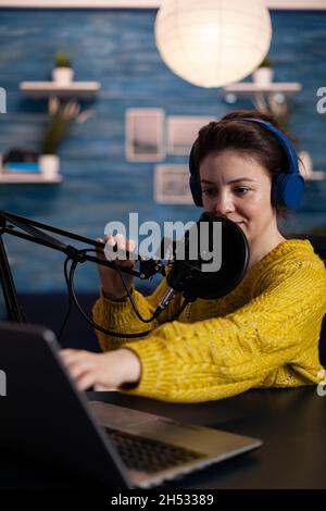 Woman vlogger looking at laptop reading listeners questions answering during online podcast at home in living room. Social media influencer recording entertainment video using production equipment Stock Photo
