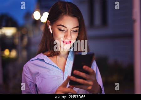 Young Caucasian businesswoman with hands free device checking social media and messages at night in city Stock Photo