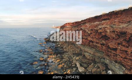 Rocky reef of shore. Shot. Top view of coastline of rocky seashore cliffs of red with holes and sticking stones. Beautiful blue sea comes and goes in Stock Photo