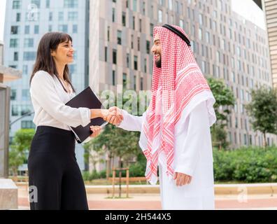 Low angle of happy businesswoman smiling and shaking hand of bearded Arab man in traditional clothes during meeting in downtown Stock Photo
