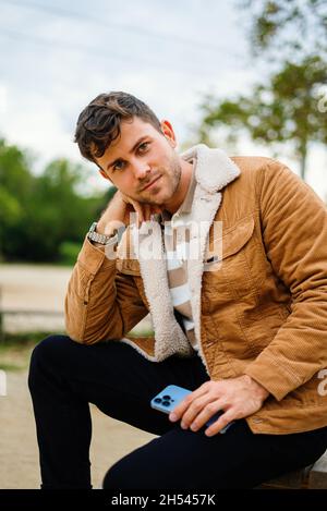 Male in warm jacket and with cellphone leaning on hand and looking at camera while sitting on blurred background of park Stock Photo