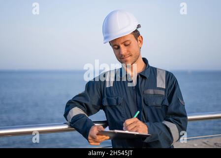 Young male logistics worker in protective helmet and workwear with clipboard signing papers while working in sea port Stock Photo