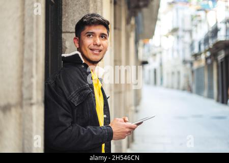 Young man standing next to a building with a tablet in his hands looking sideways and smiling. Stock Photo