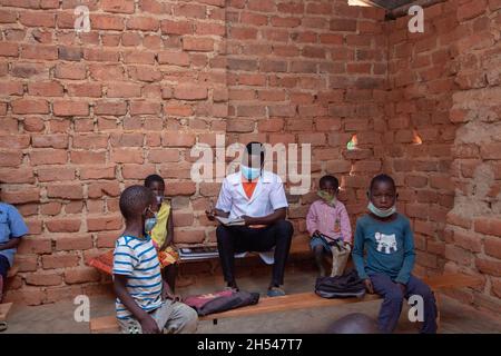 Teacher checking homework in a mud brick classroom, with students wearing a face mask and sitting apart on wooden be Stock Photo