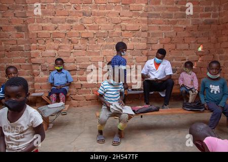 Teacher checking homework in a mud brick classroom, with students wearing a face mask and sitting apart on wooden be Stock Photo