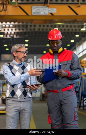 Multiracial Industrial Co-Workers Standing In A Factory And Talking About Production Process. African American Worker Writing On Clipboard. Stock Photo