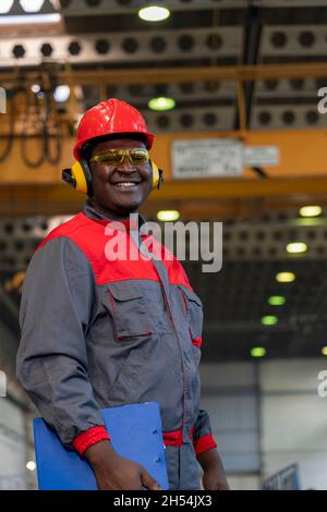 Waist Up Portrait Of Black Industrial Worker In Red Helmet, Yellow Safety Goggles, Noise Reduction Earmuffs And Work Uniform In A Factory. Stock Photo