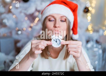 Portrait of a caucasian woman with an alarm clock in her hand and a santa hat against the background of an elegant christmas tree with a garland, in a white jacket. Selective focus Stock Photo