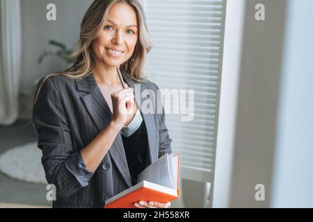 Young smiling blonde woman with long hair in stylish grey suit with diary in hands near window in the bright modern office Stock Photo