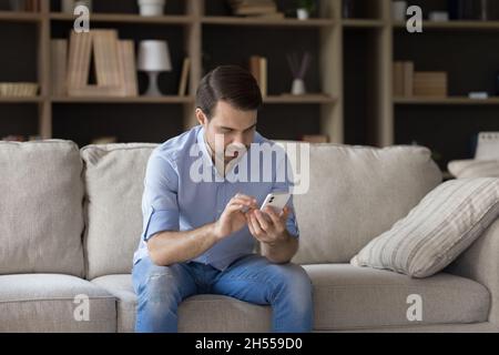 Happy millennial man using smartphone, sitting on sofa. Stock Photo