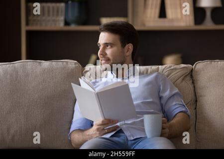 Happy dreamy relaxed young man reading book, drinking coffee. Stock Photo