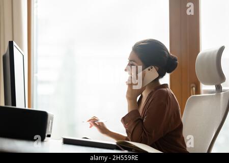 Smiling Indian business lady sit at workplace talk on smartphone Stock Photo