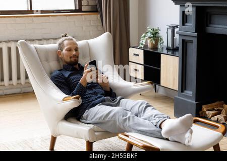 Happy relaxed young hipster man using cellphone, sitting in armchair. Stock Photo