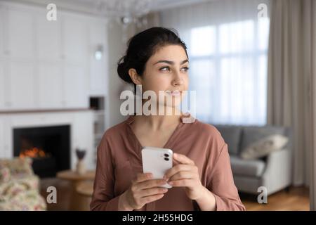 Indian woman standing indoor holds smartphone looking into distance Stock Photo