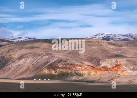 Colorful autumn landscape with colored and snowy mountains, steppe and glamping tents under construction against the backdrop of blue sky and clouds. Stock Photo