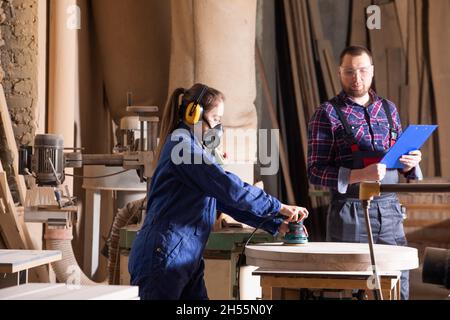 Young female apprentice polishing wood under senior colleague supervision Stock Photo