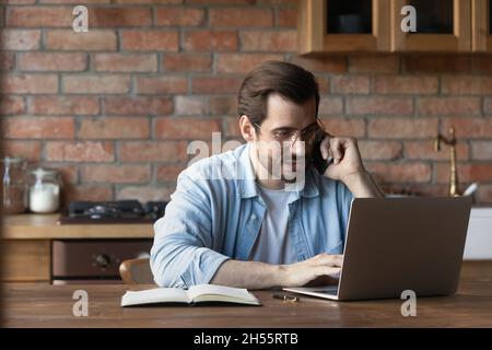 Focused millennial guy work on laptop engaged in telephone conversation Stock Photo