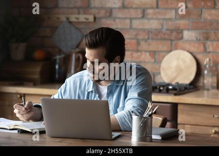 Focused guy take notes sitting by laptop at kitchen table Stock Photo