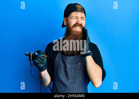 Redhead man with long beard tattoo artist wearing professional uniform and gloves touching mouth with hand with painful expression because of toothach Stock Photo