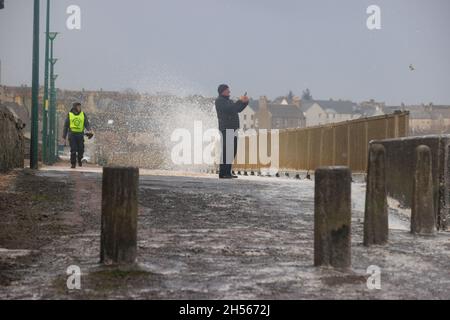 Thurso, Scotland. Nov. 7 2021. A man on a foam-covered  boardwalk takes a photograph while gale force winds send waves crashing behind him. Stock Photo