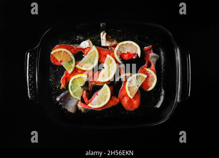 Pieces of red fish with lemon and seasoning in a glass dish on black background. Stock Photo