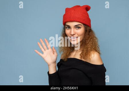 Photo of young positive cute nice brunette woman curly with sincere emotions wearing stylish black crop top and red hat isolated on blue background Stock Photo