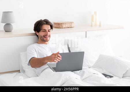 Smiling handsome young european guy with stubble play on laptop, surfing in internet, working on bed Stock Photo
