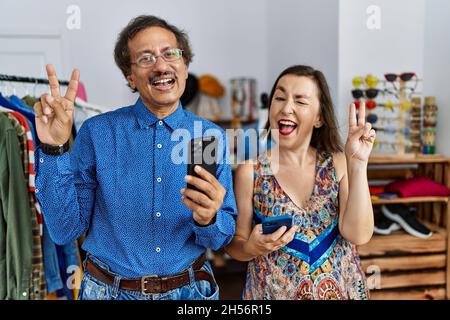 Middle age interracial couple at retail shop using smartphone smiling with happy face winking at the camera doing victory sign with fingers. number tw Stock Photo