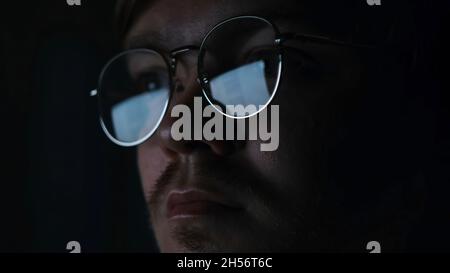 Close up of serious focused man scientist face in glasses looking at screen in the dark room. Male reading information on computer monitor that is ref Stock Photo