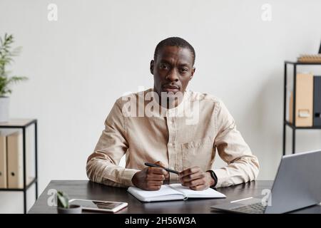 Front view portrait of black businessman looking at camera while siting at desk in minimal office, copy space Stock Photo