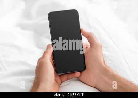 Unrecognizable hands of european young guy typing on smartphone with blank screen in bed, close up Stock Photo