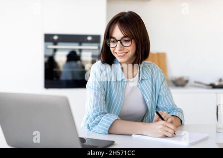 Excited freelancer lady working with laptop in kitchen interior and taking notes, writing down information Stock Photo