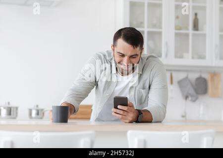 Cheerful mature european man with cup of coffee reads news and checks social networks at kitchen Stock Photo