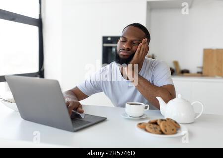 Tired black man using laptop sitting at kitchen table Stock Photo