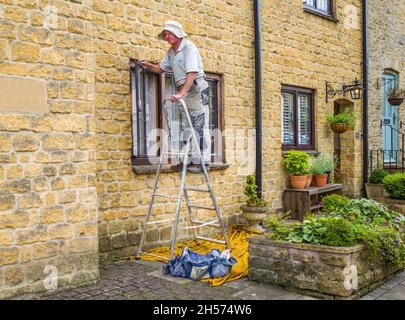 A man in overalls standing on a ladder while working on a window frame at the front of his house. Stock Photo