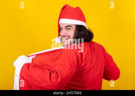 The young man is wearing Santa Claus suit. He smiles and looks towards the viewer with a mischievous look. Yellow background and copy space. Stock Photo