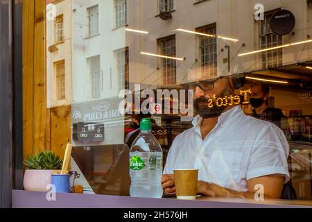 Through the window of a coffee shop a man can be seen concentrating and wearing headphones while working on a laptop computer. Stock Photo