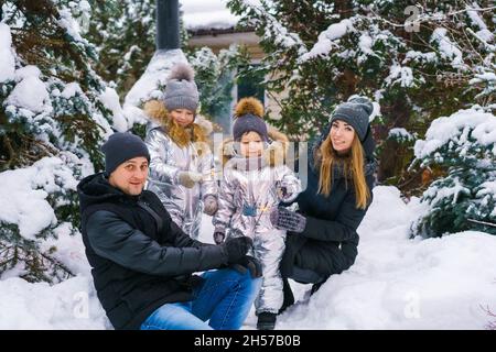 Mom with dad and son and daughter in winter in yard near snow-covered christmas tree. Falling snowflakes. Portrait of beautiful family in winter clothes near christmas tree. Family in winter forest. Stock Photo