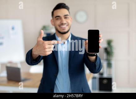 Young Arab businessman pointing at smartphone with blank screen at modern office, mockup for mobile app or website Stock Photo