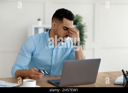 Millennial Arab entrepreneur having tired irritated eyes, overworking on laptop, exhausted from online job at office Stock Photo